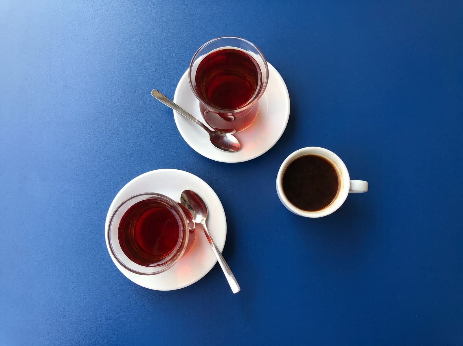 Beispielbild: Zwei Gläser Tee auf weißen Untertassen und eine Tasse Kaffee von oben auf blauer Fläche